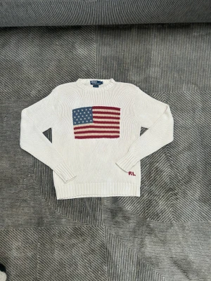 Vit stickad tröja Polo Ralph Lauren - Vit stickad tröja från Polo Ralph Lauren i 100% bomull. Framsidan har en stor amerikansk flagga i blått, rött och beige samt RL-broderi vid nederkanten. Tröjan har rund hals och långa ärmar med ribbade muddar. Perfekt för dig som gillar klassisk och cool stil.