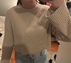 Beige croppad stickad tröja - Säljer en beige croppad stickad tröja med ribbad struktur och hög hals. Tröjan har långa ärmar och en loose passform som ger en chill vibe. Perfekt att matcha med jeans eller kjol för en snygg look.