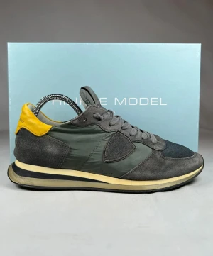 Philippe Model grå sneakers med gul detalj - Snygga sneakers från Philippe Model i grått med kontrasterande gul häl. Ovandelen är i mocka och nylon med mesh vid tån, och har snörning samt en platt sula.