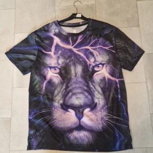 Svart t-shirt med lila lejontryck - Unik svart t-shirt från The Mountain med ett stort, detaljerat tryck av ett lila lejonansikte och blixtar över hela framsidan och baksidan. T-shirten har rund hals och korta ärmar, tillverkad i polyester. Perfekt för dig som gillar djurmotiv och vill sticka ut.
