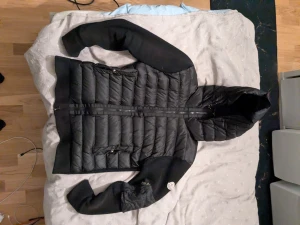 Svart Moncler pufferjacka med huva - Svart pufferjacka från Moncler med stickade ärmar och rygg, samt quiltad framsida och huva. Jackan har dragkedja framtill, två sidofickor med dragkedja och Moncler-logga på vänster ärm. Materialmix av polyester och stickad textil ger en schysst kontrast.