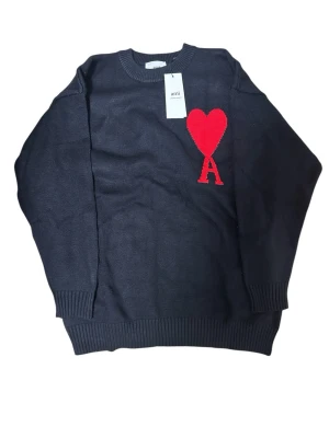 Svart stickad tröja från Ami Paris - Svart stickad tröja från Ami Paris med rund halsringning och ribbade muddar. På bröstet finns ett rött hjärta med bokstaven A, vilket ger en unik och trendig look. Perfekt för dig som gillar stilrena plagg med en twist.