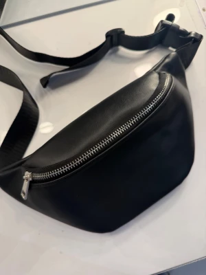 Svart magväska - Svart midjeväska som aldrig blivit använd! Den är enkel och smidig, rymmer det viktigaste och har justerbart band samt en bra dragkedja.  *Köpt men blev aldrig använd. 