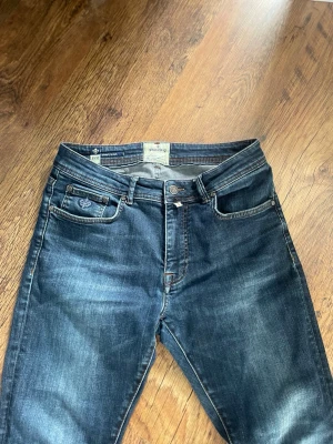 Mörkblå jeans från Morris Atelier - Snygga mörkblå jeans från Morris Atelier med klassisk femficksdesign och bruna kontrastsömmar. Jeansen har en rak passform och är tillverkade i slitstarkt denim med diskreta slitningar för en cool look. Märkespatch i skinn baktill och broderad logga på myntfickan.