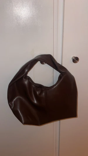 Brun axelväska i skinn - Snygg brun axelväska i mjukt skinnmaterial med rundad form och bred axelrem. Väskan har minimalistisk design utan synliga detaljer och passar perfekt för dig som gillar stilrena accessoarer. Perfekt storlek för det viktigaste.