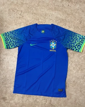 Blå Brasil Nike t-shirt med leopardmönster - Snygg blå Brasil t-shirt från Nike med coolt leopardmönster i grönt och blått på ärmarna. T-shirten har korta ärmar, Brasil-logga och Nike swoosh på bröstet. Perfekt för fotboll och sportiga dagar. Materialet är lätt polyester som andas.