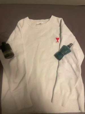 Vit stickad tröja från Ami - Vit stickad tröja från Ami med rund hals och ribbade muddar. På bröstet finns Ami:s ikoniska röda hjärta med A-bokstav broderat. Tröjan är långärmad och har en clean, stilren look som funkar till många outfits. Materialet är mjuk bomull.