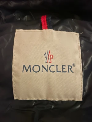 Moncler dunjacka - Snygg svart pufferjacka från Moncler med glansig finish och huva. Jackan har två fickor med dragkedja och coola Moncler-detaljer på zipparna. Quiltad design och asymmetrisk dragkedja framtill, det är en äldre modell vilket gör den väldigt unik. Perfekt för dig som vill ha både stil och värme. Köpt på vestaire för 2999, jätte fint skick. Står ingen storlek men skulle säga 170 i barn, och xs/s lite mer not s i dam. Skriv för minsta funderingar!