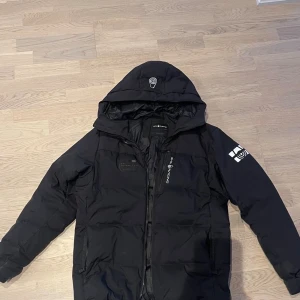 Svart Sail Racing dunjacka med huva - Svart dunjacka från Sail Racing med huva och snygga logodetaljer på bröstet och ärmarna. Jackan har dragkedja framtill, två sidofickor och är fodrad med Allied Feather + Down. Perfekt för kalla dagar och har en modern, sportig look.