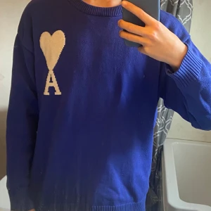 Ami Paris  - Säljer en snygg blå stickad tröja från Ami med en vit hjärtdetalj och bokstaven 'A' på bröstet. Tröjan har en rund halsringning och ribbade muddar vid ärmslut och nederkant. Perfekt för att ge en färgklick till din garderob! 💙. Köpte från en annan från plick.