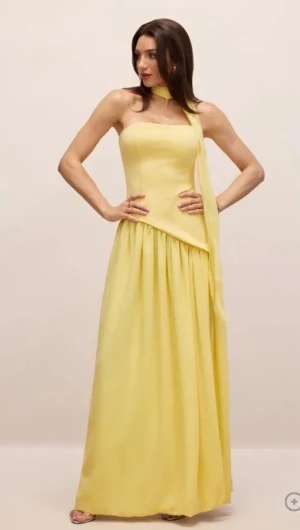 Gul långklänning med snörning - Säljer en elegant gul långklänning med axelbandslös design och asymmetrisk midja. Klänningen har ett matchande scarf.