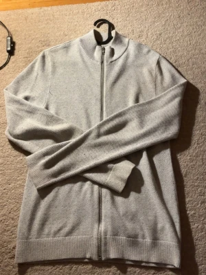 Ljusgrå kofta från Jack & Jones XS - Snygg ljusgrå kofta från Jack & Jones i storlek XS. Tröjan har ribbad krage, muddar och nederkant samt en hel dragkedja framtill. Materialet är mjukt och stickat, perfekt för lager-på-lager under höst och vinter. Enkel och clean design som funkar till många stilar.