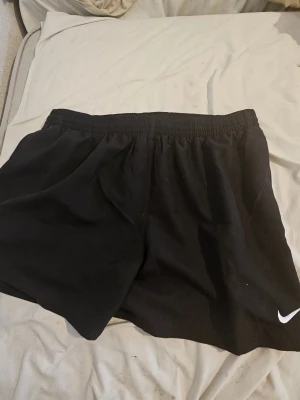 Svarta träningsshorts från Nike XXL - Svarta shorts från Nike i storlek XXL, perfekta för träning. De har en loose passform, elastisk midja och är tillverkade i lätt polyester. Klassisk Nike-logga i vitt på benet. Snygga och bekväma för gymmet eller sport.