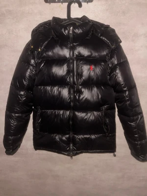 Svart dunjacka från Polo Ralph Lauren - Snygg svart dunjacka från Polo Ralph Lauren med röd broderad logga på bröstet. Jackan har huva, flera dragkedjefickor och quiltad design. Perfekt för kalla dagar och har en modern, puffad look som är riktigt trendig.