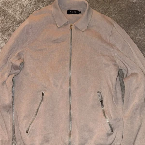 Beige stickad kofta från Riley - Säljer en beige stickad kofta från Riley med hel dragkedja framtill och två snedställda fickor med dragkedjor. Klassisk krage och ribbade muddar ger en clean look. Perfekt för lager-på-lager under höst och vår.