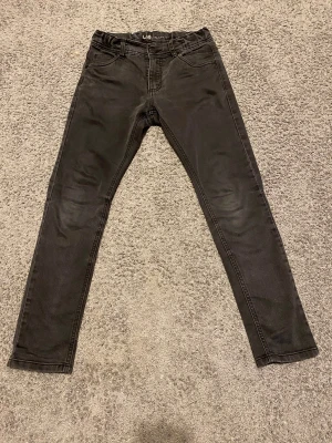 Svarta skinny jeans från LAB Industries - Svarta jeans från LAB Industries i storlek 152. Modellen är skinny med smal passform och klassisk femficksdesign. Jeansen har bälteshällor och knappstängning framtill. Materialet är mjukt och stretchigt för extra komfort.