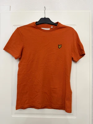Orange t-shirt från Lyle & Scott - Snygg orange t-shirt från Lyle & Scott med klassisk rund hals och korta ärmar. På bröstet sitter den ikoniska gula fågel-loggan. T-shirten är gjord i mjuk bomull och har en clean, enkel design som funkar till jeans eller shorts.