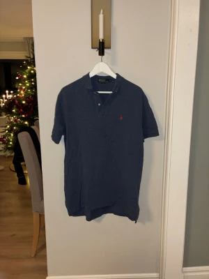  Polo Ralph Lauren - Säljer en klassisk marinblå polotopp från Polo Ralph Lauren i storlek M. Toppen har korta ärmar, krage med två knappar och den ikoniska röda Polo-loggan broderad på bröstet. Materialet är mjuk bomull som känns skön mot huden. Har två defekter ena är en fläck som borde gå att tvätta bort andra är en av knaparna som är lite konstig är du intresserad hör av dig!