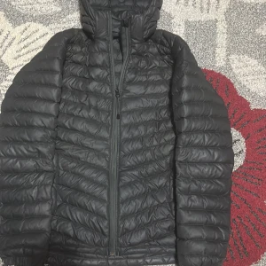 Svart pufferjacka Peak Performance - Säljer en svart pufferjacka från Peak Performance med huva och snygga quiltade detaljer. Jackan har dragkedja framtill och är tillverkad i syntetmaterial som håller dig varm. Perfekt för dig som gillar stilren och sportig look.