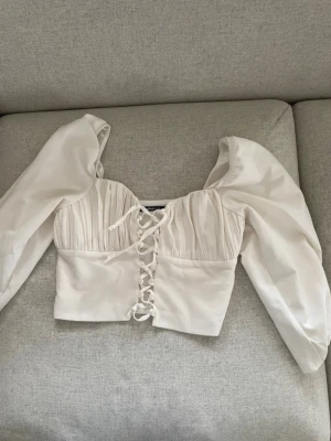 Vit snörad korsettopp från Gina Tricot. Cropped  - Supersnygg vit korsettopp från Gina Tricot med puffiga långärmar och snörning framtill. Toppen har plisserad överdel och croppad passform som ger en trendig siluett. Perfekt att styla med höga jeans eller kjol för en chic look. Det är mini-slitage som ser ut som noppror. Se sista bild. Inget man tänker på i verkligheten. Rättvis i storlek 