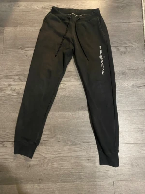 Svarta mjukisbyxor Sail Racing S - Svarta mjukisbyxor från Sail Racing i storlek S. Byxorna har snörning i midjan, ribbade muddar vid bensluten och en bakficka. Loggan är tryckt längs ena benet. Mjukt material som är perfekt för chill eller träning.