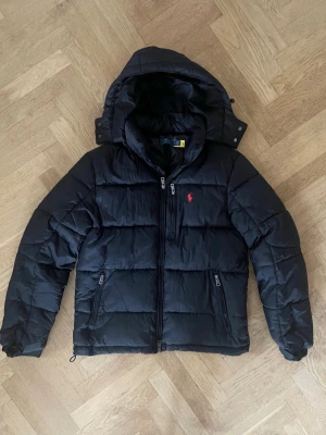 Svart pufferjacka Polo Ralph Lauren - Svart pufferjacka från Polo Ralph Lauren med huva, dragkedjor på fickor och bröst, samt den klassiska röda logon på bröstet.  Säljer den för att den blev för liten, jag har använt den i ca 1 månad och nu säljs den. 
