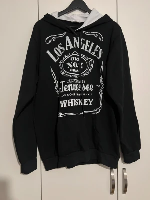 Svart hoodie Los Angeles Tennessee XL - Svart hoodie från OLK i storlek XL med vit tryckt text och motiv inspirerat av whiskeyetiketter. Kontrastfärgad huva med vit insida och svarta snören. 