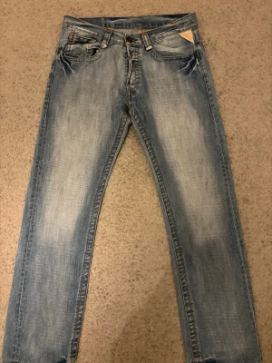 Replay ljusblå jeans med broderi - Säljer ett par ljusblå Replay jeans med snygga slitningar och broderade detaljer på bakfickorna. Klassisk femficksmodell med knappstängning och Replay-logga på läderpatch bak. Jeansen har en rak passform och är tillverkade i mjukt denimtyg. Passar både tjej och kille! Perfekt julklapp till pojkvän eller dig själv🥰