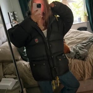 Svart dunjacka från Fjällräven  - !!!Pris kan diskuteras vid snabbt köp!!! Säljer en svart,  dunjacka från Fjällräven med klassisk röd logga på bröstet. Jackan har stora fickor med knappar, gul snörning nertill och en rejäl huva. Perfekt för kalla dagar och riktigt mysig att bära. Nypris runt 10 000 då den är gammal modell!!! Så därmed priset men skulle gå o diskutera🫶