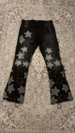 Counting Stars Stacked Slim Flare Jeans - oanvända - Säljer ett par svarta Fashion Nova-jeans med unik stjärndesign. Jag köpte dem men har aldrig hunnit använda dem då jag jobbar konstant. Därför tänker jag att någon annan får bättre nytta av dem än att de bara ligger hemma.  Jeansen är helt nya, aldrig använda och har fortfarande originallappen kvar. Modellen har distressed detaljer och stilrena gråa stjärnpatches både fram och bak.  Perfekta för dig som gillar statement-jeans och streetwear. Kan hämtas eller skickas.