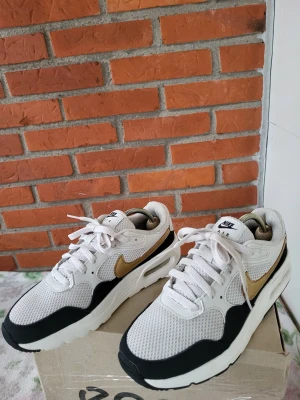 Nike Air Max sneakers vit/guld - Nike Air Max sneakers i vitt och svart med guldiga swoosh-detaljer. Ovandel i mesh och syntet, klassisk chunky sula med synlig Air-enhet. Skorna har snörning och rund tå, perfekt för dig som gillar streetstyle och sportig vibe.