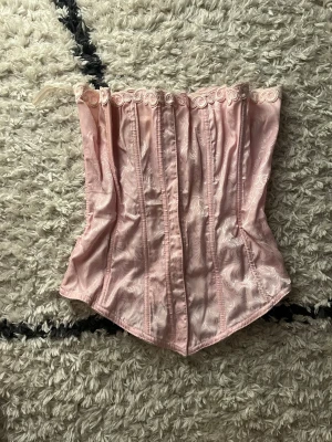 Ljusrosa korsett med spets och snörning - Säljer en ljusrosa korsett med vacker paisleyinspirerad jacquardmönstring och diskret mönster. Korsetten har snörning i ryggen för justerbar passform och är tillverkad i glansigt satinliknande material. Strokek och märke vet jag inte men skulle säga att den är i storlek S. En ring där snöret går igenom på ryggen har lossnat, för övrigt bra skick 