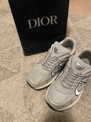 Grå Dior B30 sneakers - Säljer ett par grå Dior B30 sneakers med reflekterande detaljer och tydlig Dior-logga på sidan. Skorna har mesh och skinnpaneler, snörning och en chunky vit sula. Perfekta för dig som vill ha en lyxig och sportig vibe i din outfit. Priset kan diskuteras.