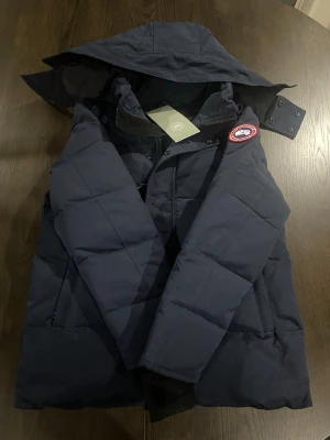 Marinblå dunjacka Canada Goose - Säljer en marinblå dunjacka från Canada Goose med huva och klassisk logotyp på ärmen. Jackan har quiltad design, dragkedja och tryckknappar framtill. Insidan är fodrad i svart och har meshficka. Perfekt för kalla dagar.