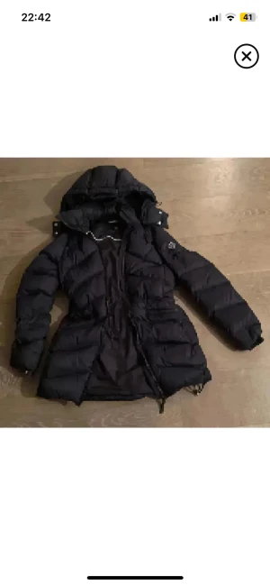 Mörkblå Gant lång pufferjacka med huva - Snygg Mörblå pufferjacka med huva och snörning i midjan. Jackan är lång, har dragkedja framtill och quiltad design som ger en cool look. Perfekt för kalla dagar och har en normal passform med långa ärmar.