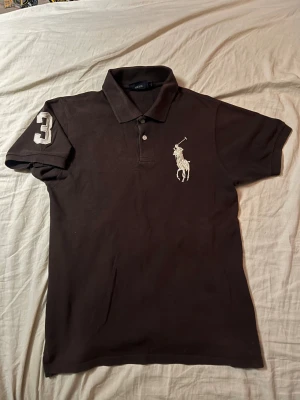Brun Polo Ralph Lauren t-shirt - Snygg brun t-shirt från Polo Ralph Lauren med klassisk polokrage och korta ärmar. Stor broderad logga i vitt på bröstet och en vit siffra 3 på ena ärmen. Tillverkad i mjuk bomull, perfekt för en chill och sportig look. Passar utmärkt på en som är M. Skriv om ni har några funderingar