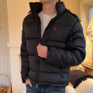 Svart pufferjacka från Polo Ralph Lauren - Svart pufferjacka från Polo Ralph Lauren med röd logga på bröstet. Jackan har hög krage, huva och två sidofickor med dragkedja. Materialet är syntet och dun, perfekt för kalla dagar. Stängs med dragkedja framtill och har en clean, modern look.