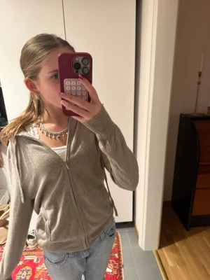 Beige kashmirkofta från Soft Goat - Mysig beige kofta från Soft Goat i kashmir. Modellen har dragkedja framtill, långa ärmar och ribbade muddar. Perfekt att slänga på sig över ett linne eller t-shirt. Enkel och stilren design som funkar året runt.