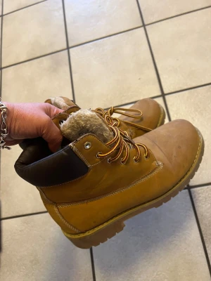 Bruna fodrade boots från Cult storlek 39 - Säljer ett par bruna boots från Cult med grov platt sula och rund tå. Skorna har snörning med gul-röda snören och är fodrade med mjuk päls på insidan för extra värme. Ovandelen är i skinn och har mörkbrunt vadderat skaft. Perfekta för kalla dagar.