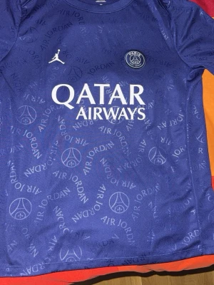 Psg training jersey  - Bilderna visar en blå PSG x Jordan fotbollströja med Qatar Airways-tryck, Air Jordan-mönster och Paris Saint-Germain-logga. Tröjan är i polyester och har Dri-Fit-teknologi, men ingen sko syns på bilderna.