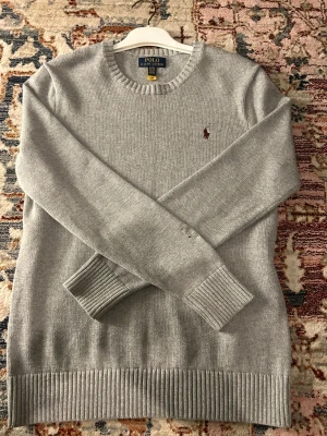 Grå stickad tröja Polo Ralph Lauren - Snygg grå stickad tröja från Polo Ralph Lauren i storlek XL. Klassisk rund hals och ribbade muddar vid ärmslut och nederkant. Broderad vinröd logga på bröstet ger en stilren touch. Perfekt för dig som gillar enkel och tidlös design.