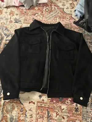 Svart overshirt med dragkedja - Svart overshirt med klassisk krage, två stora bröstfickor med lock och sidofickor. Jackan har en silverfärgad dragkedja framtill och vita knappar vid ärmsluten. Materialet känns mjukt och har en matt finish, perfekt för lager-på-lager.