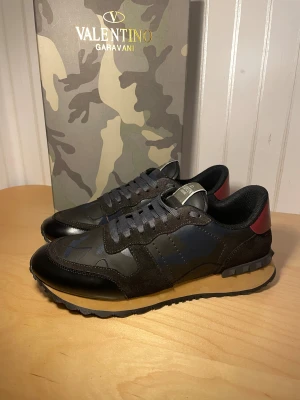 Valentino Garavani Rockrunner sneakers - Snygga Valentino Garavani Rockrunner sneakers med mörk camouflage-mönster i svart och blått, detaljer i rött på hälen och beige sula. Skorna har snörning, platt sula och är tillverkade i skinn och mocka med coola nitar på hälen. Perfekt för dig som gillar exklusiv streetstyle.