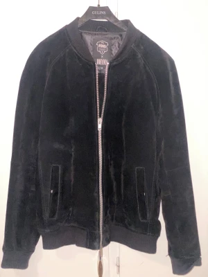 Svart teddy bomberjacka King x Jofama - Svart teddy bomberjacka från King x Jofama med mjuk yta och silverfärgad dragkedja framtill. Jackan har ribbade muddar vid ärmslut och nederkant, samt två fickor på framsidan. Perfekt för dig som gillar stilren och bekväm streetwear.