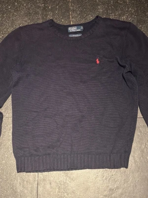 Mörkblå stickad tröja Polo Ralph Lauren - Mörkblå stickad tröja från Polo Ralph Lauren med klassisk röd broderad logga på bröstet. Tröjan har rund halsringning, ribbade muddar och är tillverkad i 100% bomull. Perfekt för dig som gillar stilrena och tidlösa plagg.