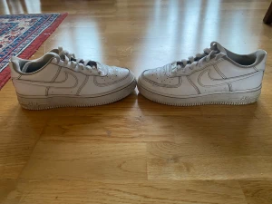 Nike Air Force 1 vita sneakers - Klassiska Nike Air Force 1 sneakers i helvitt skinn med rund tå och platt sula. Skorna har snörning och den ikoniska swoosh-loggan på sidorna. Perfekta för dig som gillar en clean och tidlös look. Passar till det mesta och är riktigt bekväma.