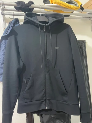 Hugo boss kofta  - Svart hoodie från BOSS med stilren logga på bröstet, hel dragkedja och snörning i huvan. Tröjan har två fickor framtill och är gjord i mjuk bomullsmix. Perfekt för chill dagar och streetwear vibes.