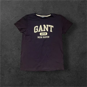 Mörkblå GANT t-shirt med tryck - Snygg mörkblå t-shirt från GANT i storlek S med klassiskt tryck 'GANT 1949 NEW HAVEN' på bröstet. T-shirten har rund hals och korta ärmar, tillverkad i mjuk bomull som är skön mot huden. Perfekt för dig som gillar sportig och stilren design.