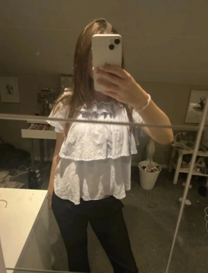 blus med volanger - Säljer en söt vit blus med dubbla volanger och korta ärmar. Blusen har en luftig passform och är tillverkad i ett lätt bomullstyg. Perfekt för dig som gillar romantiska detaljer och vill ha en fräsch look. Volangerna ger en extra touch och gör toppen unik.