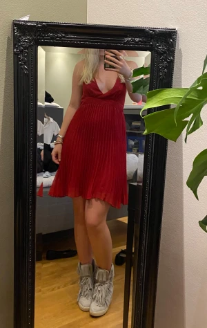 Röd plisserad kortklänning med öppen rygg - Säljer en snygg röd kortklänning med plisserad kjol och djup v-ringning. Klänningen har tunna axelband som knyts i nacken och en öppen rygg för en cool och ungdomlig look. Perfekt för dig som vill sticka ut med en färgstark och trendig stil.